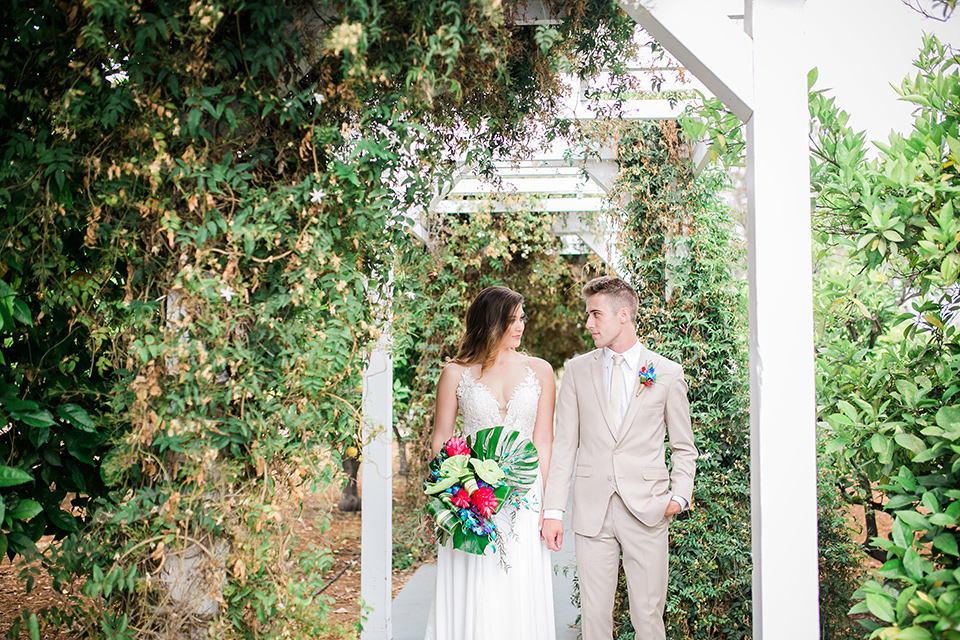 bride in a boho style gown with a bright colored floral crown and the groom in a tan suit with a tan long tie walking outside