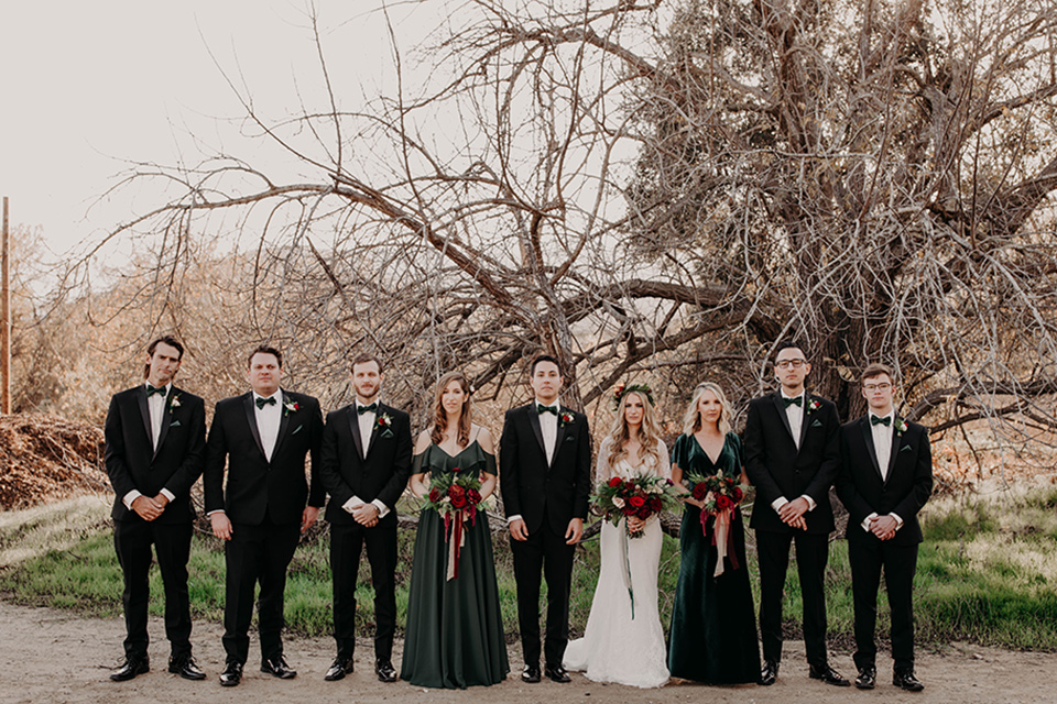 Highway-West-Vacations-bridal-party-all-lined-up-bridesmaids-in-dark-green-dresses-groomsmen-in-black-tuxedos-with-green-velvet-bow-ties-bride-in-a-lace-bohemian-dress-with-sleeves-and-floral-crown-groom-in-a-black-tuxedo