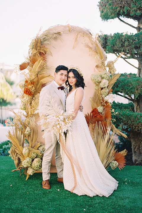  bride in a cream colored bohemian inspired gown with a gold crown, groom in a tan suit with a burgundy velvet bow tie, with a bohemian style ceremony arch 