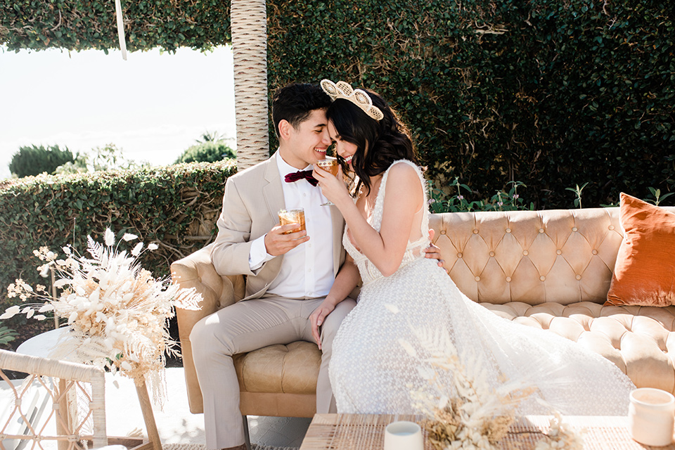  bride in a cream colored bohemian inspired gown with a gold crown, groom in a tan suit with a burgundy velvet bow tie on the couch 