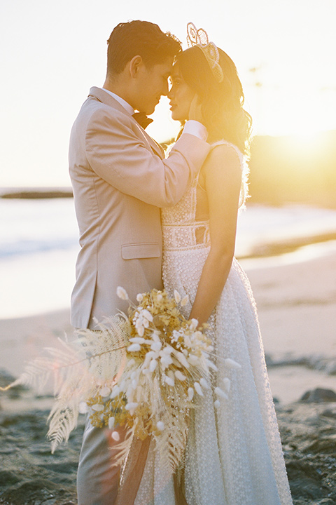  bride in a cream colored bohemian inspired gown with a gold crown, groom in a tan suit with a burgundy velvet bow tie, at the beach 