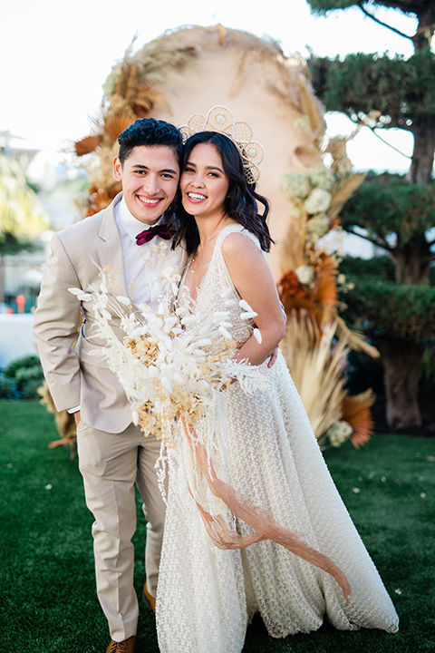  bride in a cream colored bohemian inspired gown with a gold crown, groom in a tan suit with a burgundy velvet bow tie, with a bohemian style ceremony arch 