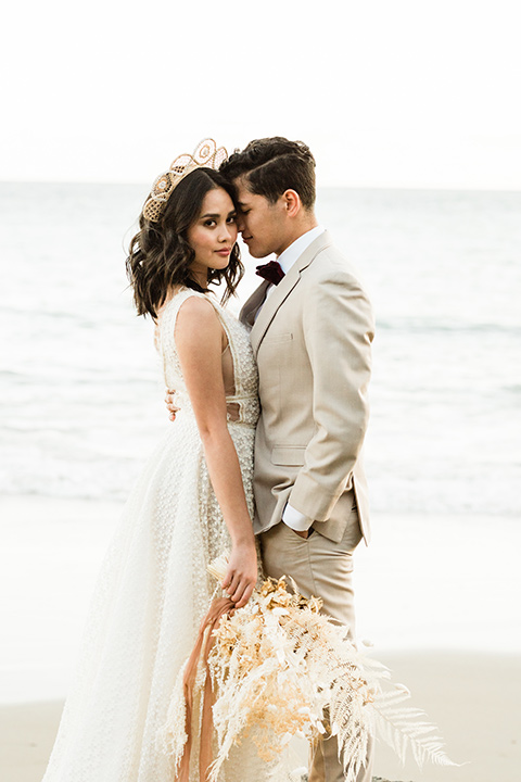  bride in a cream colored bohemian inspired gown with a gold crown, groom in a tan suit with a burgundy velvet bow tie, at the beach 