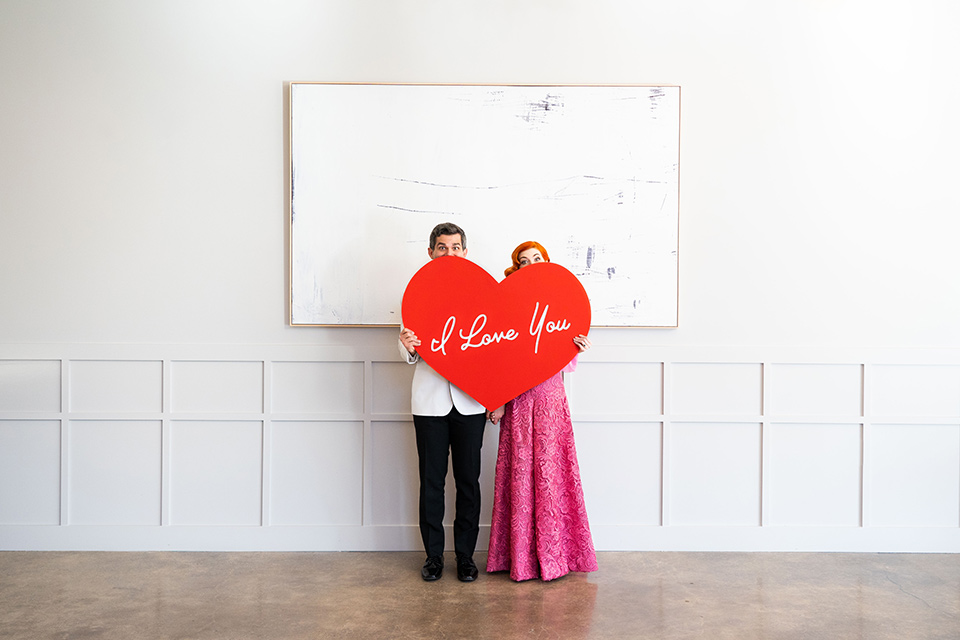 I-love-lucy-shoot-bride-and-groom-behind-heart-sign-bride-in-a-pink-wide-legged-pant-and-top-with-red-hair-and-red-lipstick-groom-in-a-white-and-black-tuxedo