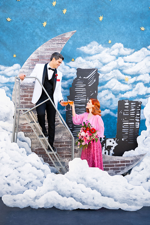 I-love-lucy-shoot-bride-and-groom-cheersing-by-moon-bride-in-a-pink-wide-legged-pant-and-top-with-red-hair-and-red-lipstick-groom-in-a-white-and-black-tuxedo