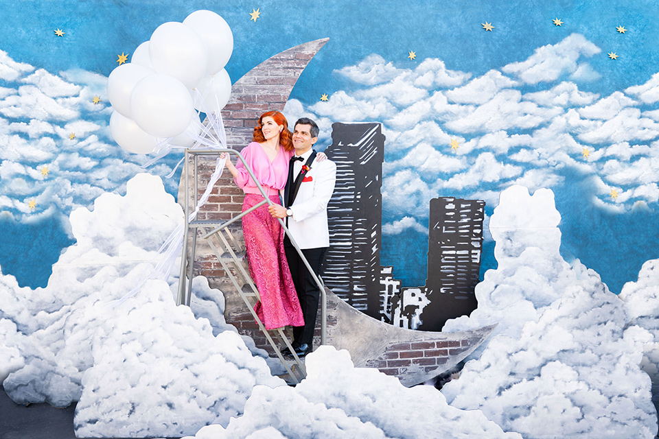 I-love-lucy-shoot-bride-and-groom-holding-balloons-by-moon-backdro-bride-in-a-pink-wide-legged-pant-and-top-with-red-hair-and-red-lipstick-groom-in-a-white-and-black-tuxedo