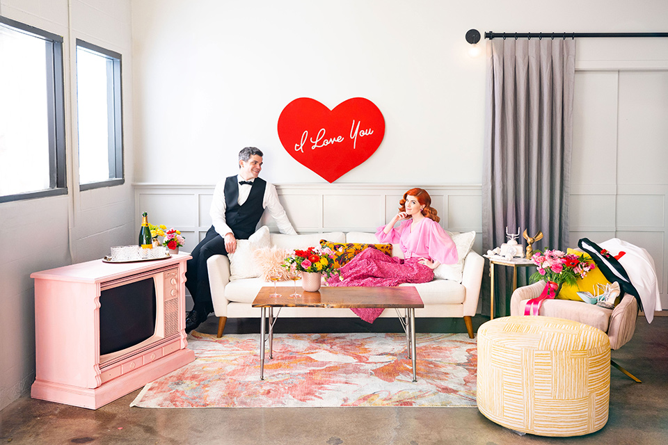 I-love-lucy-shoot-bride-and-groom-on-the-couch-bride-in-a-pink-wide-legged-pant-and-top-with-red-hair-and-red-lipstick-groom-in-a-white-and-black-tuxedo