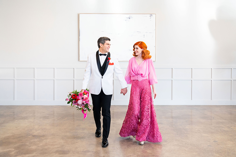 I-love-lucy-shoot-bride-and-groom-walking-towards-camera-bride-in-a-pink-wide-legged-pant-and-top-with-red-hair-and-red-lipstick-groom-in-a-white-and-black-tuxedo