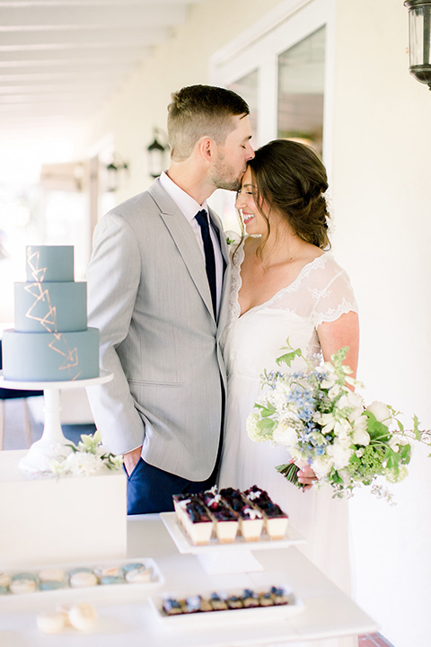 bride and groom by cake, bride in a flowing gown with a lace bodice and cap sleeves, the groom in a light grey suit coat with dark blue pants and a long tie