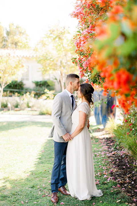 bride and groom by flowers, bride in a flowing gown with a lace bodice and cap sleeves, the groom in a light grey suit coat with dark blue pants and a long tie