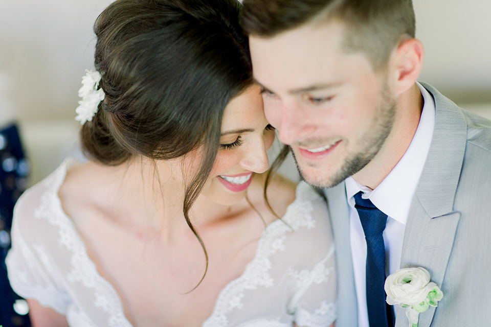  bride and groom close up sitting on the couch, bride in a flowing gown with a lace bodice and cap sleeves, the groom in a light grey suit coat with dark blue pants and a long tie 