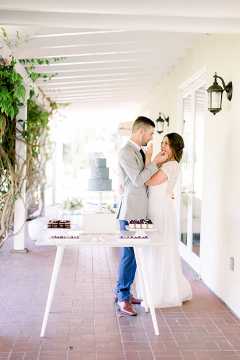  bride and groom by dessert table, bride in a flowing gown with a lace bodice and cap sleeves, the groom in a light grey suit coat with dark blue pants and a long tie 