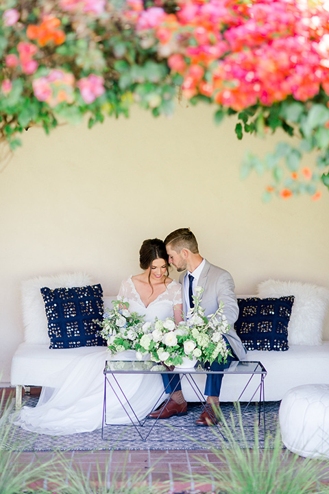  bride and groom on couch, bride in a flowing gown with a lace bodice and cap sleeves, the groom in a light grey suit coat with dark blue pants and a long tie 