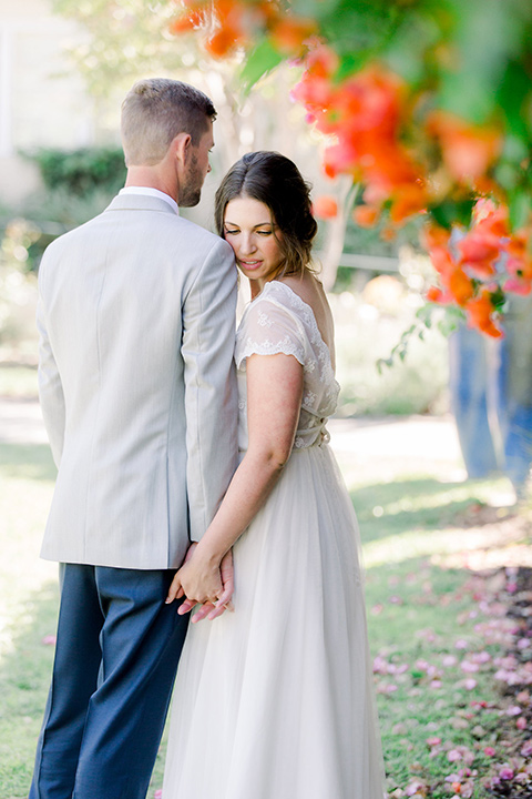  bride and groom in garden, bride in a flowing gown with a lace bodice and cap sleeves, the groom in a light grey suit coat with dark blue pants and a long tie 