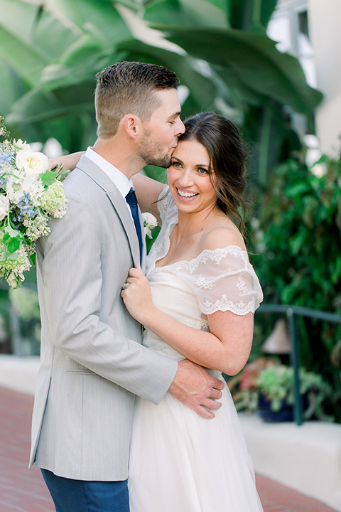  bride and groom hugging, bride in a flowing gown with a lace bodice and cap sleeves, the groom in a light grey suit coat with dark blue pants and a long tie 