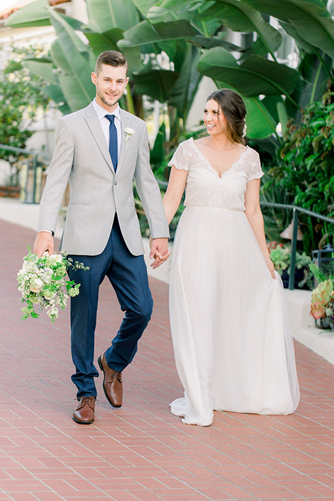 bride and groom walking, bride in a flowing gown with a lace bodice and cap sleeves, the groom in a light grey suit coat with dark blue pants and a long tie