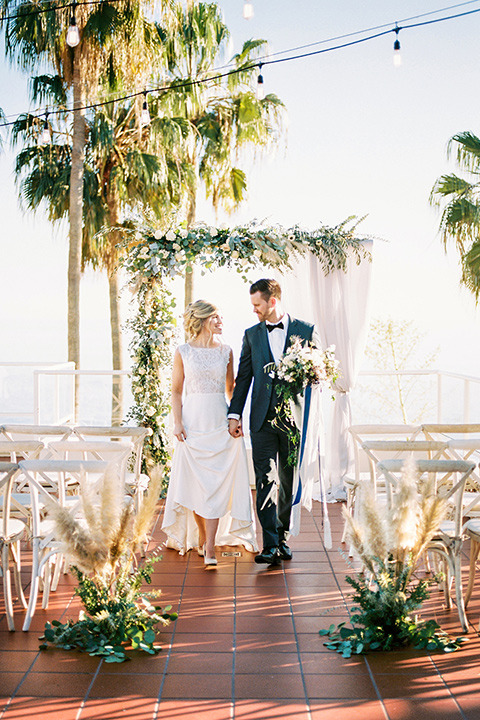 The-Inn-at-Laguna-Beach-bride-and-groom-walking-after-ceremony-bride-in-a-form-fitting-gown-with-a-high-slit-and-high-neckline-groom-in-a-grey-blue-suit-with-a-matching-bow-tie