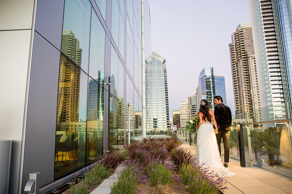 Intercontinental-elopement-shoot-bride-and-groom-walking-by-building-groom-in-velvet-tuxedo-with-black-ants-and-a-black-bow-tie-bride-in-a-flowing-gown-with-straps-and-lace