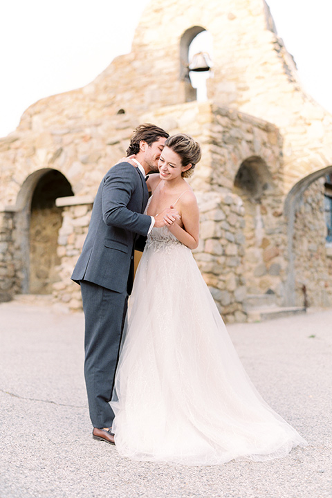 italian-style-wedding-bride-and-groom-dancing--by-stone-wall-bride-in-a-flowing-gown-with-straps-groom-in-a-grey-tuxedo-with-black-bow-tie