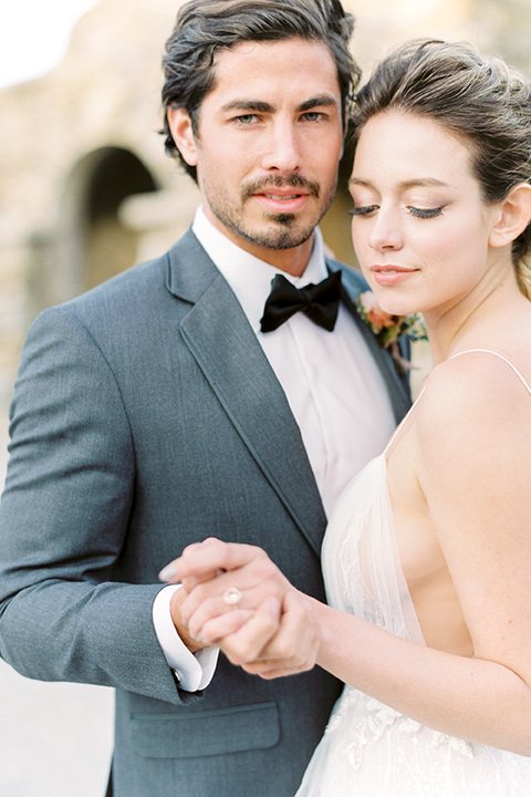 italian-style-wedding-groom-looking-at-camera-holding-bride-close-bride-in-a-flowing-gown-with-straps-and-her-hair-up-groom-in-a-grey-tuxedo