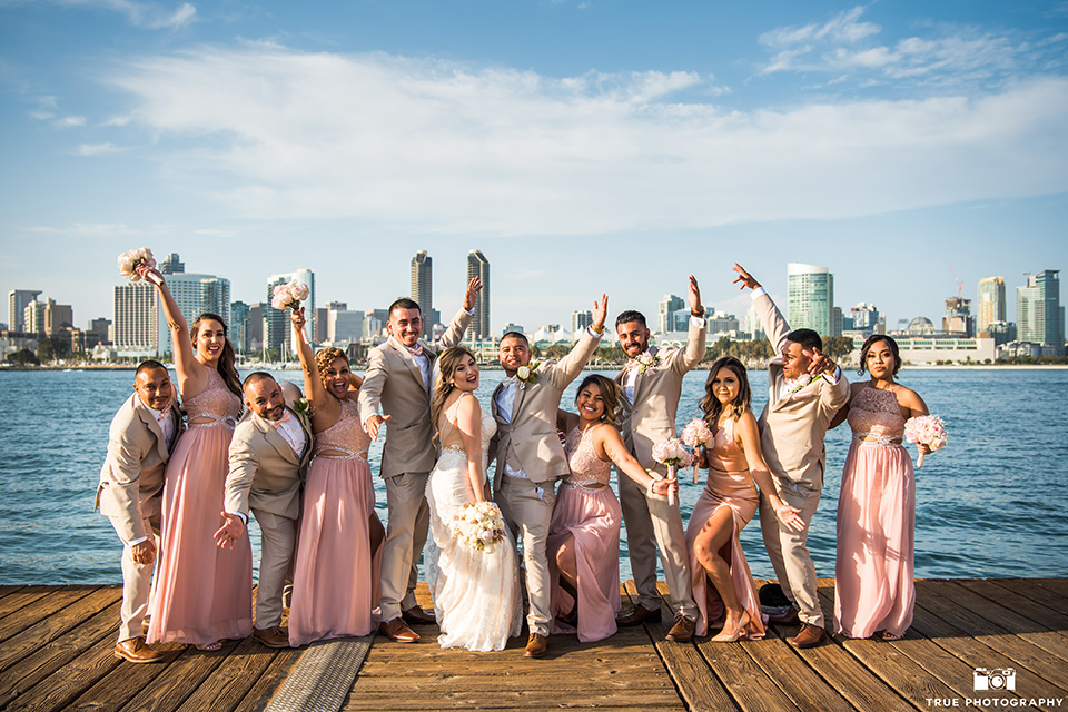 bridal party poses by the water with the bridesmaids wear blush pink long dresses and the groomsmen wearing tan notch lapel suits