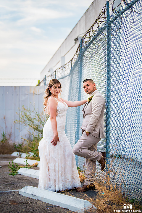 bride and groom pose in front of fence with the bride wearing an ivory lace dress and the groom is wearing a tan suit with a pink bow-tie