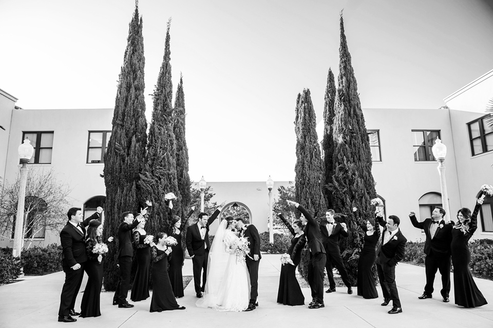 black and white photo of bridal party cheering for couple with bridesmaids in black lace gowns groomsmen in black tuxedos bride in a lace and tulle gown with a high neckline and sleeves groom in a traditional black tuxedo