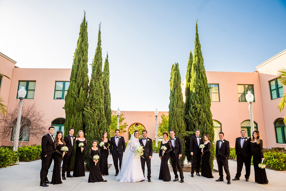 Admiral Kidd Club bridal party standing looking at camera bridesmaids in black lace gowns groomsmen in black tuxedos bride in a lace and tulle gown with a high neckline and sleeves groom in a traditional black tuxedo