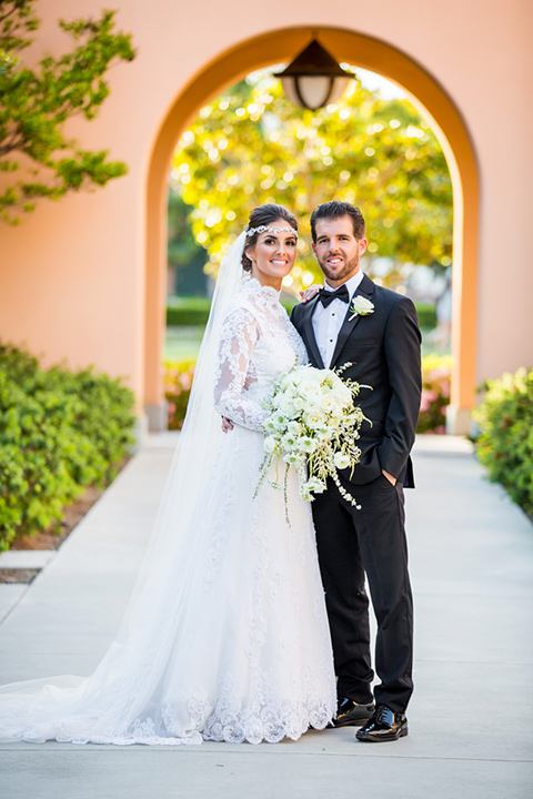 Admiral Kidd Club bride and groom standing under archway bride in a lace and tulle gown with long sleeves and high neckline groom in a traditional black tuxedo with black bow tie