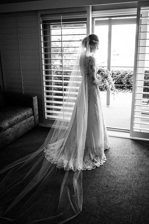 black and white photo of the bride looking out a window in a lace and tulle gown with long sleeves and high neckline groom in a traditional black tuxedo with black bow tie 