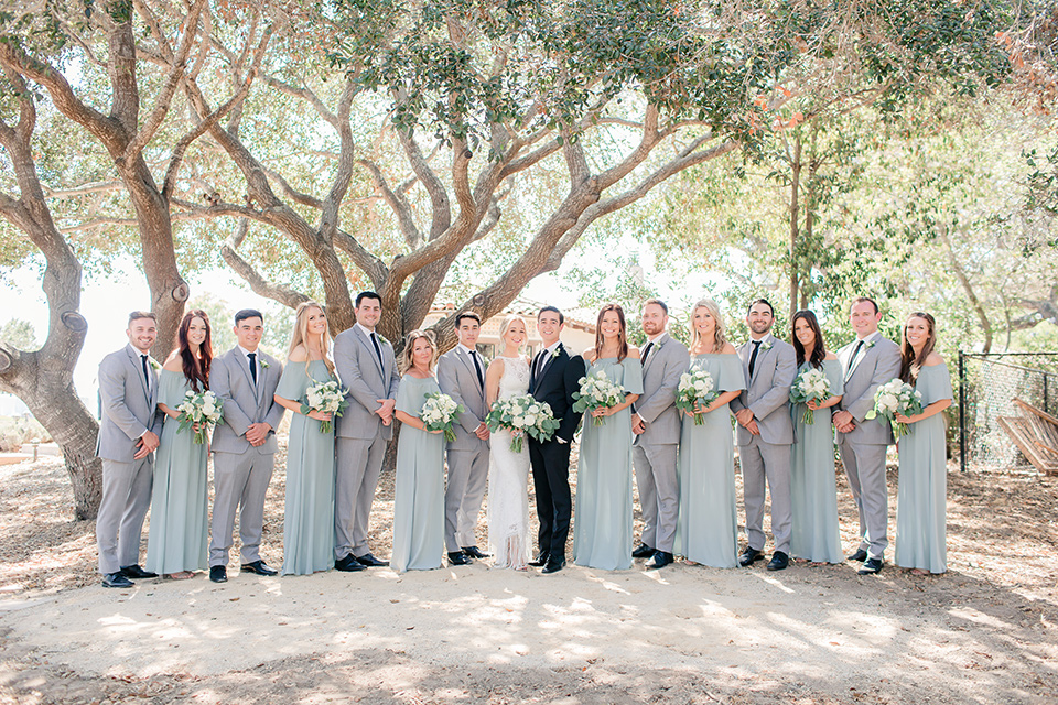 Arroyo-Grande-Wedding-bridal-party-in-a-line-bridesmaids-in-seafoam-light-green-dresses-groomsmen-in-light-grey-suits-bride-in-a-boho-style-gown-with-a-keyhole-back-and-high-neckline-and-fringe-white-the-groom-wore-a-traditional-black-tuxedo-and-black-long-tie