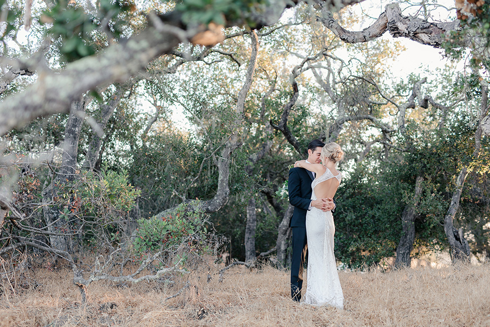 Arroyo-Grande-Wedding-bride-and-groom-hugging-under-tree-bride-in-a-boho-style-gown-with-a-keyhole-back-and-high-neckline-and-fringe-white-the-groom-wore-a-traditional-black-tuxedo-and-black-long-tie