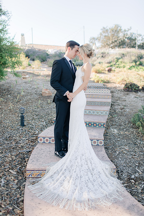 Arroyo-Grande-Wedding-bride-and-groom-on-steps-touching-heads-bride-in-a-boho-style-gown-with-a-keyhole-back-and-high-neckline-and-fringe-white-the-groom-wore-a-traditional-black-tuxedo-and-black-long-tie