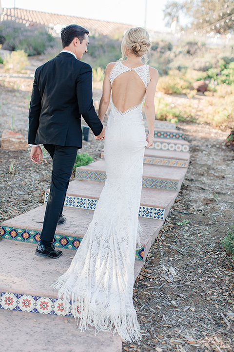 Arroyo-Grande-Wedding-bride-and-groom-walking-away-bride-in-a-boho-style-gown-with-a-keyhole-back-and-high-neckline-and-fringe-white-the-groom-wore-a-traditional-black-tuxedo-and-black-long-tie