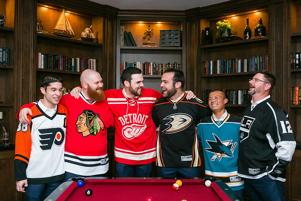 groom and groomsmen pose in front of pool table with arms around each other and wearing hockey jerseys