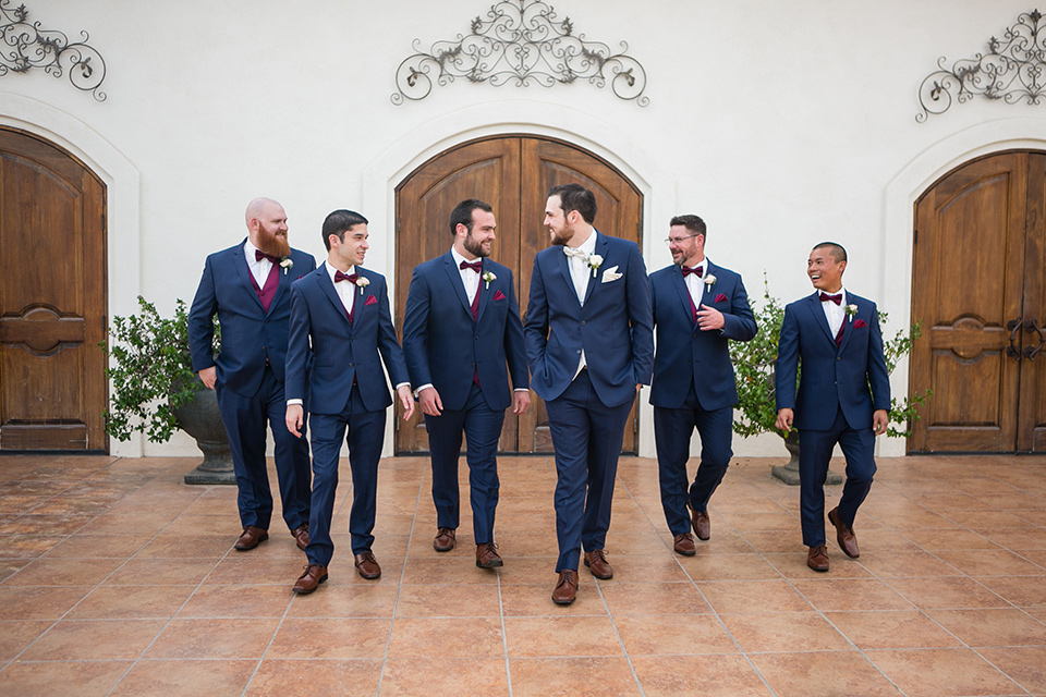 groom walks with the groomsmen in a blue suit with a white vest while groomsmen wear burgundy vests