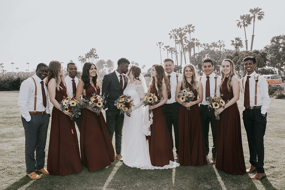 coronado-yacht-club-wedding-bride-and-groom-kissing-with-bridal-party-all-around-them-bridesmaids-in-red-dresses-groomsmen-in-a-casual-look-with-just-grey-pants-white-shirts-and-red-long-ties-bride-in-a-lace-form-fitting-gown-groom-in-a-charcoal-grey-suit-with-red-tie