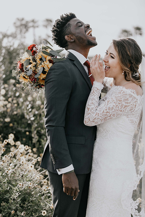 coronado-yacht-club-wedding-bride-and-groom-laughing-bride-in-a-lace-gown-with-sleeves-and-off-the-shoulder-detailing-groom-in-a-charcoal-grey-suit-and-deep-red-long-tie