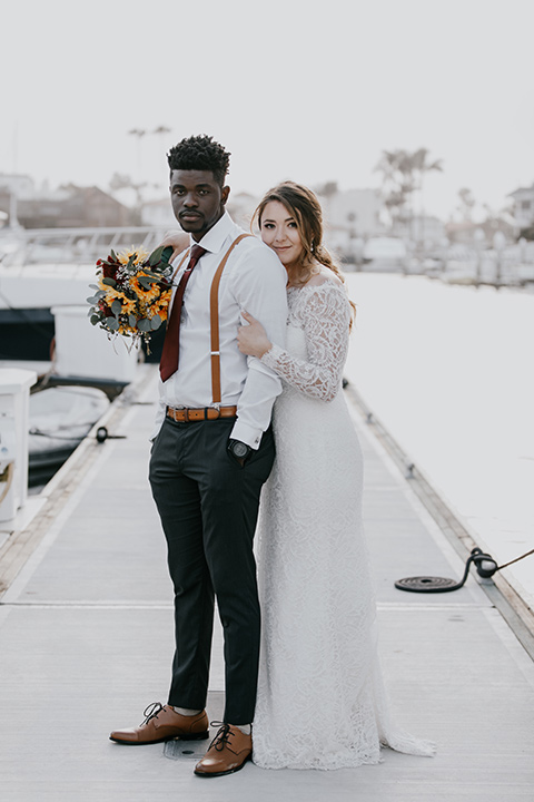 coronado-yacht-club-wedding-bride-and-groom-standing-at-the-marina-bride-in-a-lace-gown-with-sleeves-and-off-the-shoulder-detailing-groom-in-a-charcoal-grey-suit-and-deep-red-long-tie