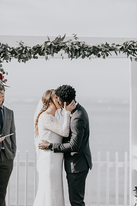 coronado-yacht-club-wedding-ceremony-kisss-bride-in-a-lace-dress-with-sleeves-and-groom-in-a-charcoal-suit-with-a-red-tie