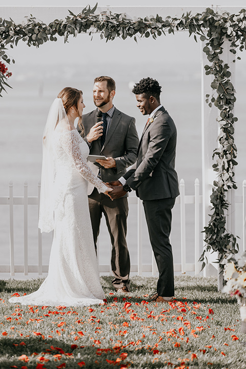 coronado-yacht-club-wedding-ceremony-bride-in-a-lace-gown-with-sleeves-and-off-the-shoulder-detailing-groom-in-a-charcoal-grey-suit-and-deep-red-long-tie