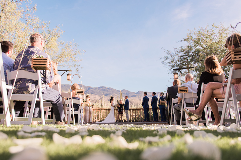 tanque-verde-ranch-arizona-wedding-ceremony-low-angle-bridesmaids-in-blush-toned-dresses-and-groomsmen-in-dark-blue-suits-the-bride-in-a-white-formfitting-gown-with-thin-straps-and-the-groom-and-in-grey-suit-pants-and-a-blue-suit-coat