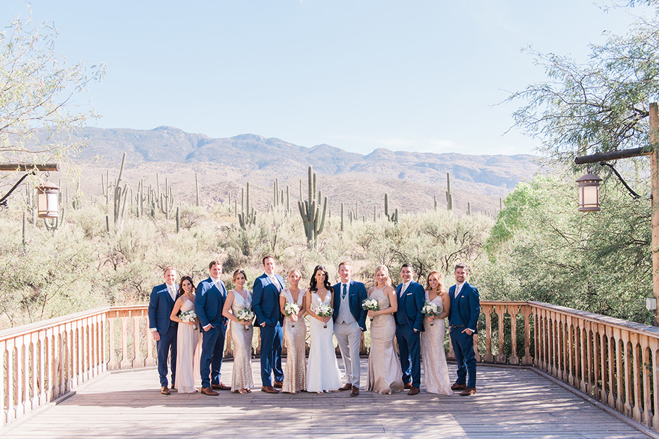 tanque-verde-ranch-arizona-wedding-bridalparty-in-a-line-bridesmaids-in-blush-toned-dresses-and-groomsmen-in-dark-blue-suits-the-bride-in-a-white-formfitting-gown-with-thin-straps-and-the-groom-and-in-grey-suit-pants-and-a-blue-suit-coat