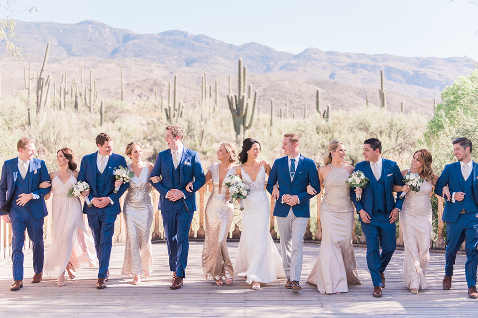 tanque-verde-ranch-arizona-wedding-groomsmen-and-bridesmaids-walking-with-couple-bridesmaids-in-blush-toned-dresses-and-groomsmen-in-dark-blue-suits-the-bride-in-a-white-formfitting-gown-with-thin-straps-and-the-groom-and-in-grey-suit-pants-and-a-blue-suit-coat