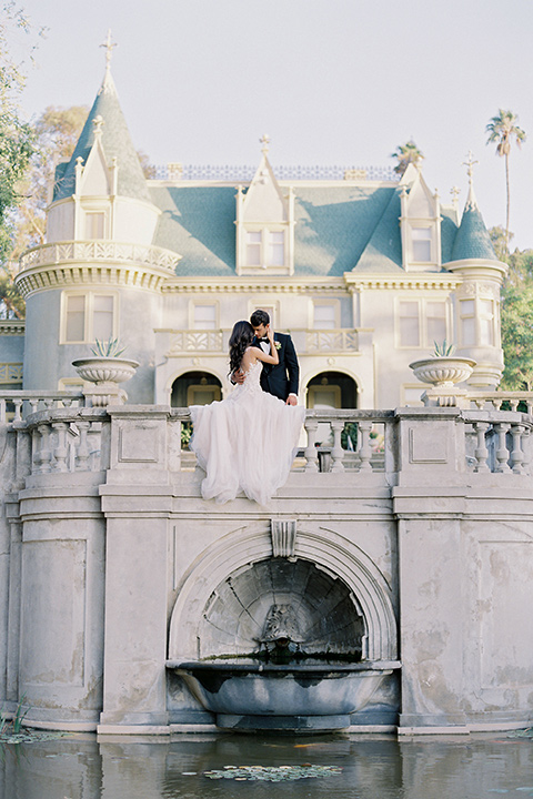 Kimberly-crest-house-shoot-bride-and-groom-sitting-on-venue-bridge-with-pond-groom-in-a-traditional-black-tuxedo-with-white-shirt-and-black-bow-tie-bride-in-a-modern-princess-inspired-flowing-white-gown-with-a-full-skirt