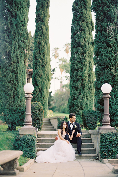 Kimberly-crest-house-shoot-bride-and-groom-sitting-groom-in-a-black-tuxedo-with-white-shirt-and-black-bow-tie-bride-in-a-flowing-white-gown-with-a-full-skirt