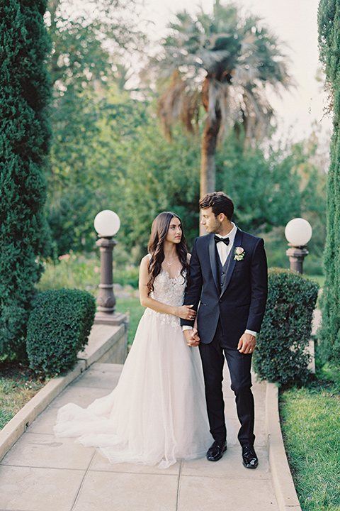 Kimberly-crest-house-shoot-bride-and-groom-walking-down-path-groom-in-a-traditional-black-tuxedo-with-white-shirt-and-black-bow-tie-bride-in-a-modern-princess-inspired-flowing-white-gown-with-a-full-skirt