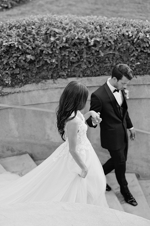 Kimberly-crest-house-shoot-bride-and-groom-walking-down-the-stairs-groom-in-a-black-tuxedo-with-white-shirt-and-black-bow-tie-bride-in-a-flowing-white-gown-with-a-full-skirt