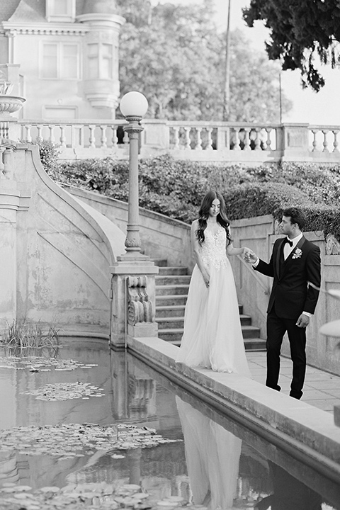 Kimberly-crest-house-shoot-groom-holding-brides-hand-by-pond-groom-in-a-black-tuxedo-with-white-shirt-and-black-bow-tie-bride-in-a-flowing-white-gown-with-a-full-skirt