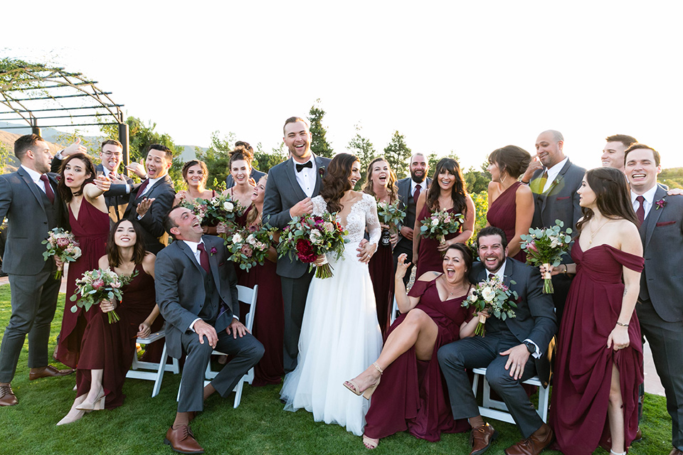  bride in a white lace gown with a full skirt and long lace sleeves and her hair in a finger wave and the groom in a charcoal tuxedo with a black bow tie and the groomsmen in charcoal tuxedo and bridesmaids in burgundy gowns